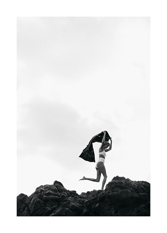 - Zwart-wit foto van een vrouw in een witte bikini, die over rotsen rent en een handdoek boven haar hoofd houdt