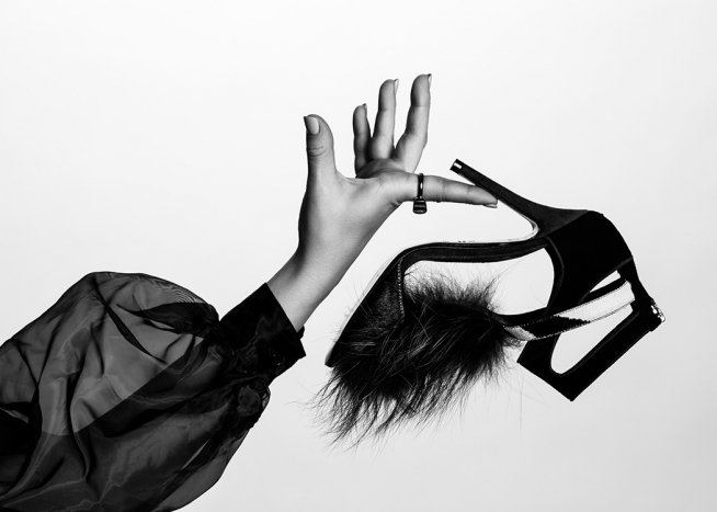  – Zwart-wit foto van een schoen met hoge hak en bont op de neus, vastgehouden door een vrouw in een transparante blouse met pofmouw