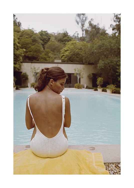  – Foto van een vrouw in een wit badpak, die op een handdoek voor een zwembad zit en een wit badpak draagt