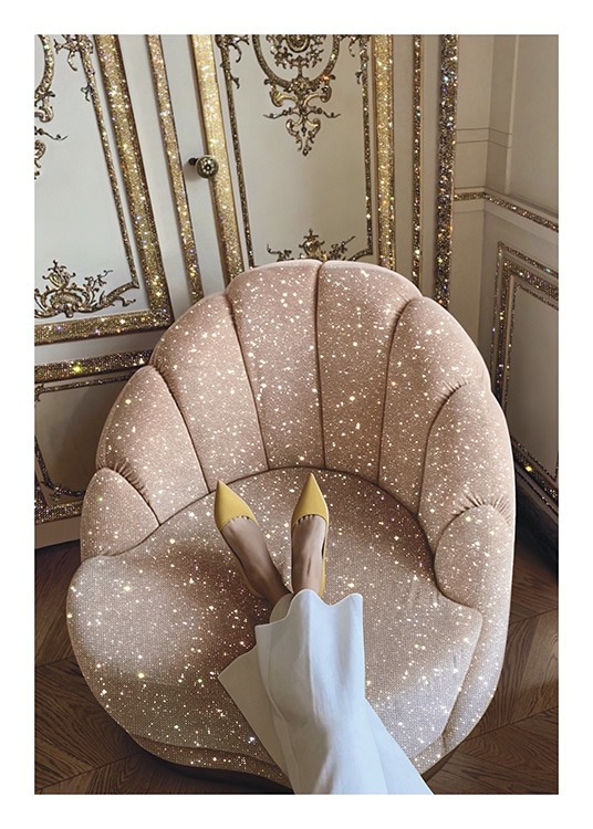  – Foto van een roze, fonkelende stoel voor een gouden en witte muur, met een vrouw die haar voeten op de stoel laat rusten