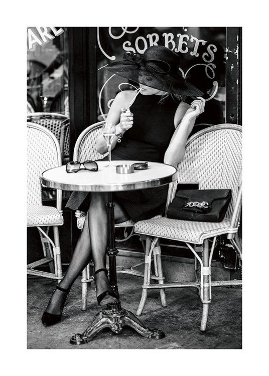  – Zwart-wit foto van een vrouw die buiten zit bij een café, terwijl ze een hoed draagt en een sigaret opsteekt